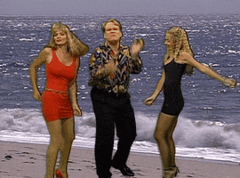 Celebrity gif. Andy Richter wears slacks and a patterned shirt as he sings earnestly. Two women in party dresses dance beside him while waves crash on a beach in the background.