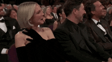 Oscars 2024 GIF. Carey Mulligan, seated at the Oscars, giggles, absentmindedly tucking her hair and blotting her lips, as she enjoys a joke.