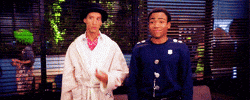 TV gif. Wearing Halloween costumes, Danny Pudi as Abed and Donald Glover as Troy on Community do the wave with their arms and give each other a smug high five.