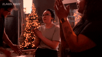 Reality TV gif. In a studio with a lit Christmas tree, three contestants on Blown Away clap their hands and snap fingers, and the middle one says "Merry frickin' Christmas," which appears as text.