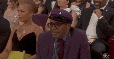 Celebrity gif. Spike Lee wears a purple suit and beret at the Academy Awards where he leans forward with a skeptical expression on his face.
