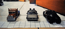 Stop motion gif. Toy Batmobile, gray car from Ghostbusters, and Optimus Prime truck race each other along a tiled floor, with the Batmobile leading the way as they zoom around a corner.