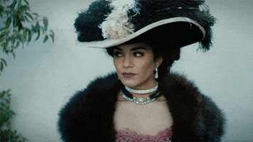 TV gif. Vanessa Hudgens as Mata Hari on Drunk History wears a large hat with extravagant feathers and a fur shawl over a dress. She purses her lips like she’s kissing the air, but in a mysterious and sexy manner.