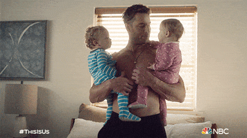 TV gif. Justin Hartley as Kevin on This is Us. He's shirtless at home and carries two babies, one in each arm, and is bouncing them on his hip.