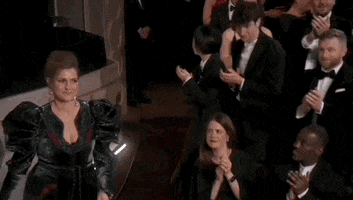 Oscars 2024 GIF. Holly Waddington briskly walks down the aisle to accept her Oscar, throwing a brief wave of casual confidence at her applause.