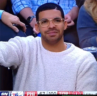 Celebrity gif. Wearing gold-rimmed glasses and a gray sweater, Drake seems to be sitting courtside at a basketball game. He claps, then stands up, claps more emphatically, and cheers.