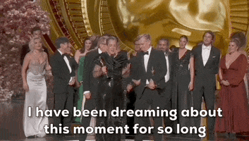 Oscars 2024 GIF. Emma Thomas holds the trophy for Best Picture for Oppenheimer while the cast and crew stand proudly behind her. She says, "I have been dreaming about this moment for so long."