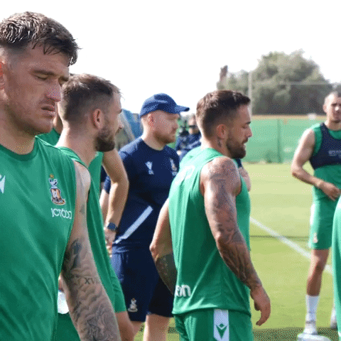 Sports gif. Andy Cook of St. Patrick's Athletic is standing with his teammates during warm ups. He stops and looks at us, scrunching his face like he's totally over this practice. 