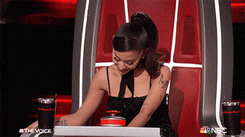 TV gif. Ariana Grande on The Voice sits in the big red chair. She looks down, shaking her head and putting her hand up to stop something.