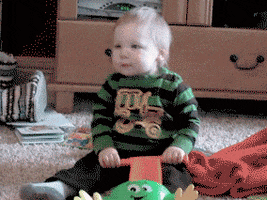 Video gif. A baby sits in a living room holding a push toy, dancing and wiggling around, moving his shoulders. 