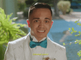 Celebrity gif. Yung Bae wears a white textured tuxedo with a teal bow tie. He bounces while looking anxious but trying to cover it with a smile. Text, "Internal panic."