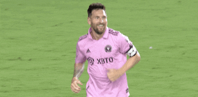 Sports gif. Soccer player Leo Messi wearing a pink Adidas jersey smiles with an open mouth, holding out his hands and running across the field toward someone.