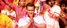 Celebrity gif. Salman Khan looks at us with a smirk and holds his hands up close to his mouth in a prayer position. People walk around him, celebrating and waving flags.