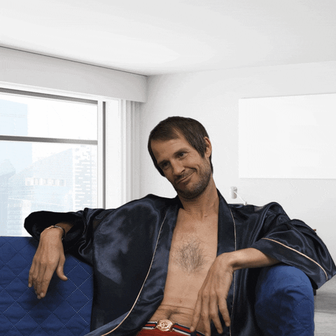 Video gif. Man with side parted bangs sits luxuriously in an open silk blue robe with no shirt underneath. He gives himself a high five and looks at someone smugly while pointing a finger gun at them.