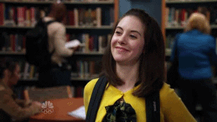 TV gif. Alison Brie as Annie Edison. She grins dorkily, leaning back on a table and begins to wave at someone but realizes the person isn't waving at her and she awkwardly touches her hair instead.