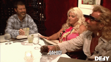 Reality TV gif. Duane in Dog the Bounty Hunter lifts a glass to cheers a man across the table as Beth looks on. Text, "Salud!"