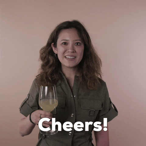 Reaction gif. A Disabled Vietnamese-American woman hemorrhagic stroke survivor with left-sided hemiplegia, brown sun-bleached hair styled in waves raises a glass of white wine to us, saying, "Cheers!"