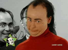 TV gif. The camera zooms in on Tim Heidecker as Spagett wearing a red turtleneck and messy mullet. He tilts his head into his double chin and stares up with a funny look on his face like he’s judging you. 