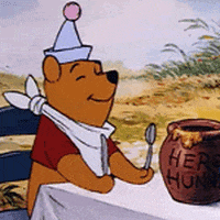 Disney gif. Winnie the Pooh sits at an outdoor table with a pot of "hero hunny" in front of him. He wears a party hat and napkin bib, and holds a spoon and knife as he bounces happily from side to side in anticipation.