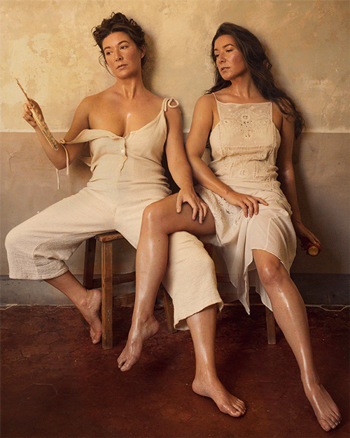 Video gif. Two women sit next to each other with overlapped legs, dewy with sweat; the left one waves a fan at her face, making the other woman's hair billow slightly.