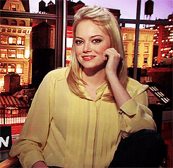 Celebrity gif. Sitting in a chair in front of a city backdrop, Emma Stone smiles and points her finger at us.