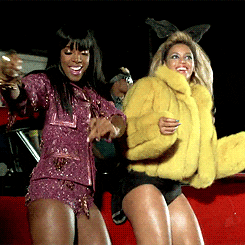 Music video gif. From the video for "Party", Beyonce wears a fluffy yellow jacket and cat ears while she does a swaying dance with a woman in a sequined maroon outfit.