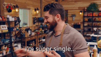 TV gif. A man wearing a kitchen apron on "The Great American Recipe" makes his way around the kitchen turning to look at us with a shy but open smile. Text, "I'm super single... super single."