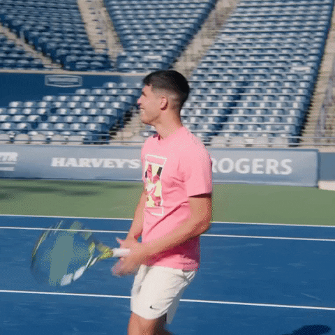 Video gif. Carlos Alcaraz on a tennis court leans back and lifts his arms in the air, holding a racket in one hand to a giant empty stadium behind him. He turns towards us and laughs as he walks back towards the net like he's goofing around. 