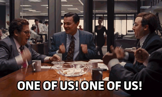 Movie gif. Leonardo Dicaprio as Jordan, Jonah Hill as Donnie, and several other characters in Wolf of Wall Street sit around a meeting table, banging their fists in unison onto the table. They excitedly cheer together, One of us! One of us!