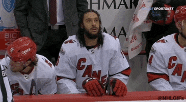 Sports gif. Hockey player on the Carolina Hurricanes sits on the bench, bouncing his shoulders up and down and looking focused like he's trying to loosen up, tossing his long hair back over his shoulders as another player looks over at what he's doing with a sassy eyebrow raise.