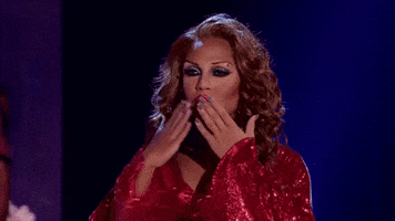 Reality TV gif. Glammed up contestant on RuPaul's Drag Race blows a kiss to the crowd and begins to strut away in her shimmering red long sleeve bikini top.