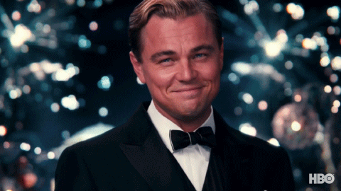 Movie gif. Leonardo DiCaprio as Jay in the Great Gatsby dons a tux and slick hair. He smiles and tips his head forward as he lifts a glass of wine, fireworks erupting in the background.