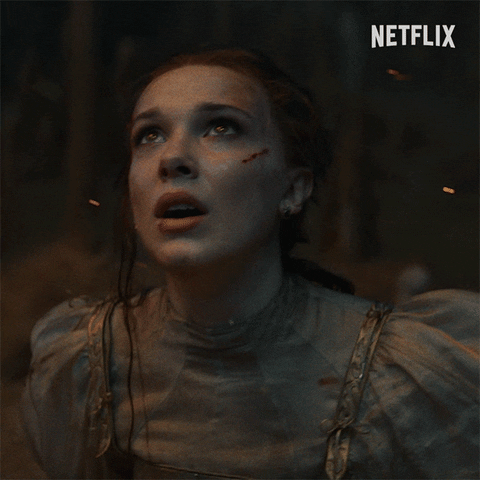 Movie gif. Millie Bobby Brown as Princess Elodie in Damsel. She's in a forest and embers fly all around her. She has cuts on her face and she pants as she looks around, horrified, staring up at the sky with wide eyes.