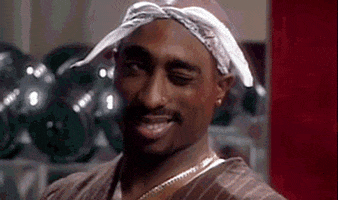 Celebrity gif. Tupac wears a white bandana tied around his head as he smiles and winks to the side.