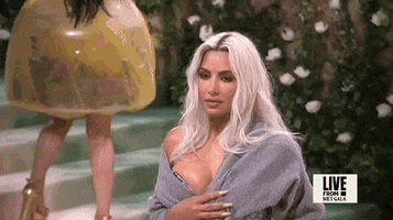 Met Gala 2024 gif. Kim Kardashian wearing a silver Maison Margiela gown and gray cardigan bolero, poses on the stairs, adjusting her sweater.