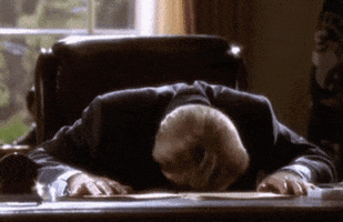 Video gif. Man sits at an office desk and bangs his head down on the desk in frustration.