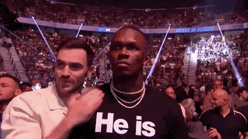 Video gif. Boxer Adonis Stevenson stands up in the crowd at a UFC fight and looks and stares something down with an intimidating gaze fixed on one point. A man leans in to whisper something in Stevenson's ear, but he doesn't waver. Behind him is a stadium of fans and spotlights. 