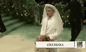 Met Gala 2024 gif. Cole Escola wears a Thom Browne monotone ivory-colored jacket, collared shirt, and bowtie. Escola swiftly turns and walks away then turns back, showing their tea-length ivory pencil skirt  with a slit at the back, ivory tights and pumps. They are wearing a circular brimmed hat with a sheer ivory veil hanging down the back. 