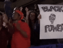 TV gif. We pan across a crowd of black people dancing and cheering in the bleachers while one person holds up a sign saying, "Black power," and another sign saying, "Black people," on Chappelles Show.