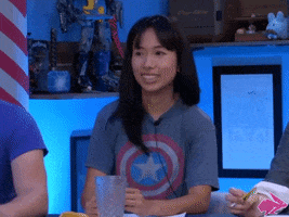 Video gif. A woman wearing a Captain America t-shirt hears good news and she sits up in her chair while using both thumbs to point at herself in pride.