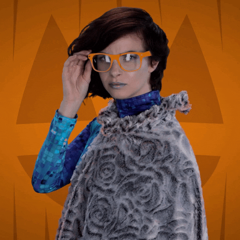 Video gif. Woman dressed in a blue turtleneck with a gray rose-patterned shawl pulls down her orange framed glasses, looking critically at us over her shoulder, then tapping her glasses back onto her face.