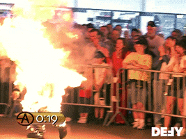 TV gif. Criss Angel walks around with his entire back on fire, a large crowd spectating him from behind metal barricades. A gold timer with white text in the bottom left counts from nineteen seconds to twenty-four seconds.