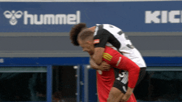 Sports gif. Video clip of two soccer players, one in a red jersey and one in a black and white jersey. The man in the black and white jersey is riding piggyback as he leans his head down to give the other player a big hug around the neck. 