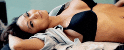Video gif. Woman laying on a table with her shirt unbuttoned and we see her black bra.