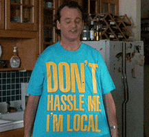 Movie gif. Bill Murray as Bob in What About Bob, holds his large t shirt spread out by the corners so we can read it. He nods up in down in a perfect loop as he smiles. The shirt says, "Don't hassle me I'm local."