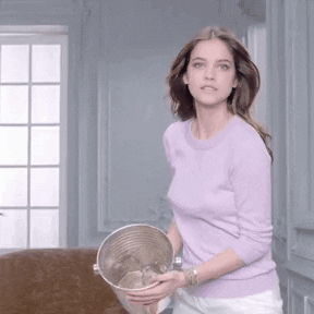 Video gif. Woman steps forward holding a shiny silver pail of water which splashes out at us in slow motion.