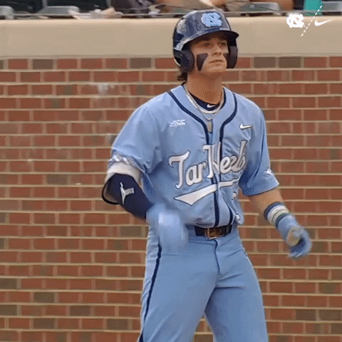 Sports gif. Colby Wilkerson, a baseball player from the University of North Carolina, is on the field wearing a batter's helmet as he stands and claps then points at someone.