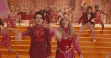 Video gif. Musicians Taylor swift and Brandon dance in colorful mod clothing in the "Me!" music video.