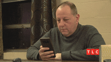 Reality TV gif. David from 90 Day Fiancé sits at a table holding his phone, looking straight-faced but surprised, raising his eyebrows and glancing side to side.|100%x100%