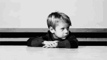 Video gif. A black and white video of a young boy waiting with his arms resting on a table, drumming his fingers.